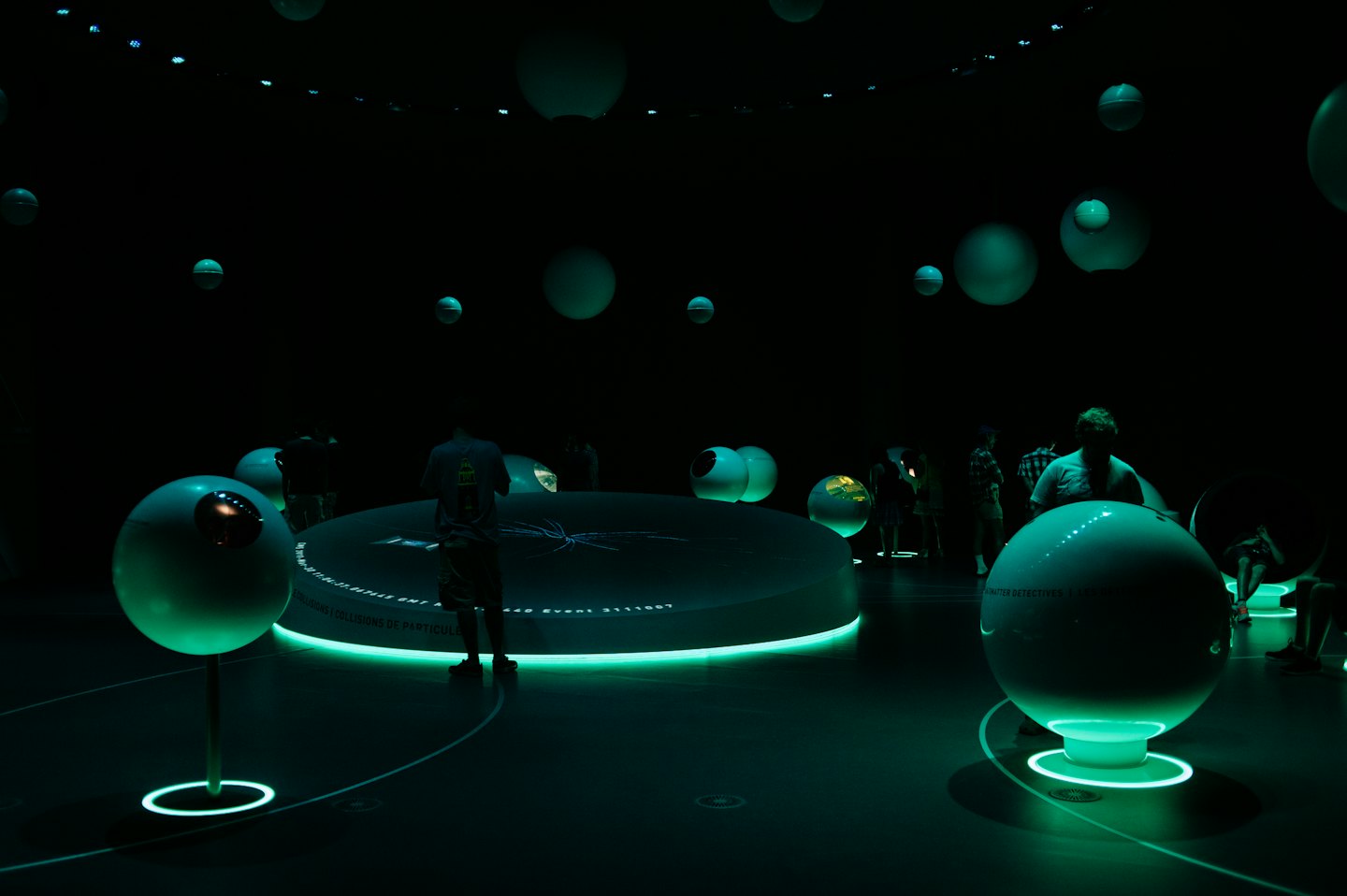 Visitors interact with glowing teal spheres in a dark science exhibit.